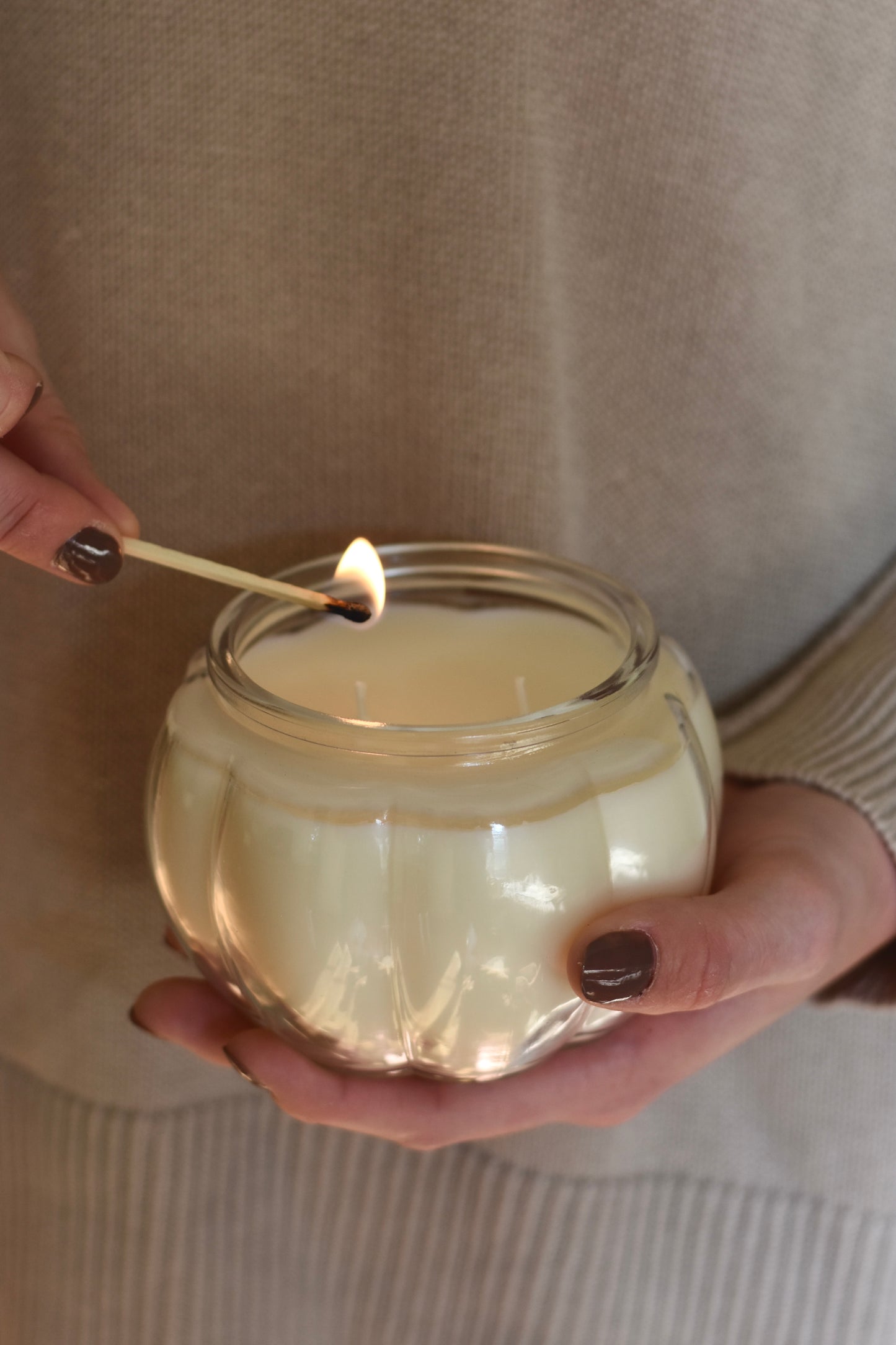 Person lighting a candle in a glass jar with a beige sweater background