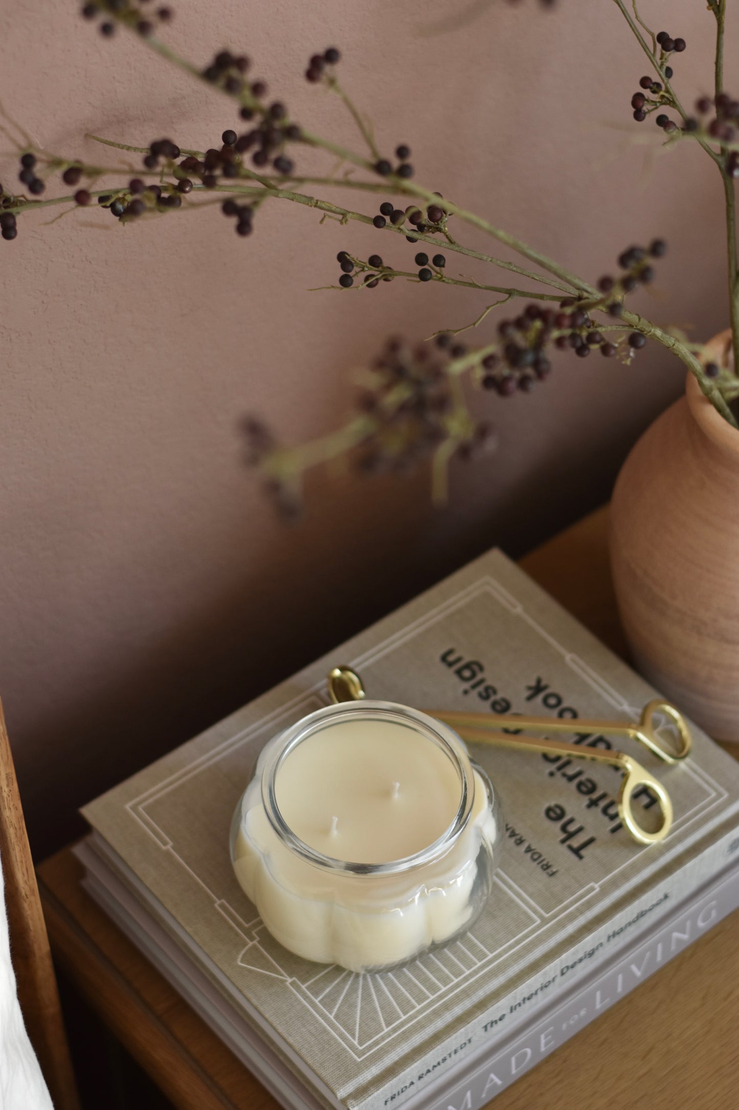 Candle in a decorative container on a book with a pair of wick trimmers, surrounded by dried plants.