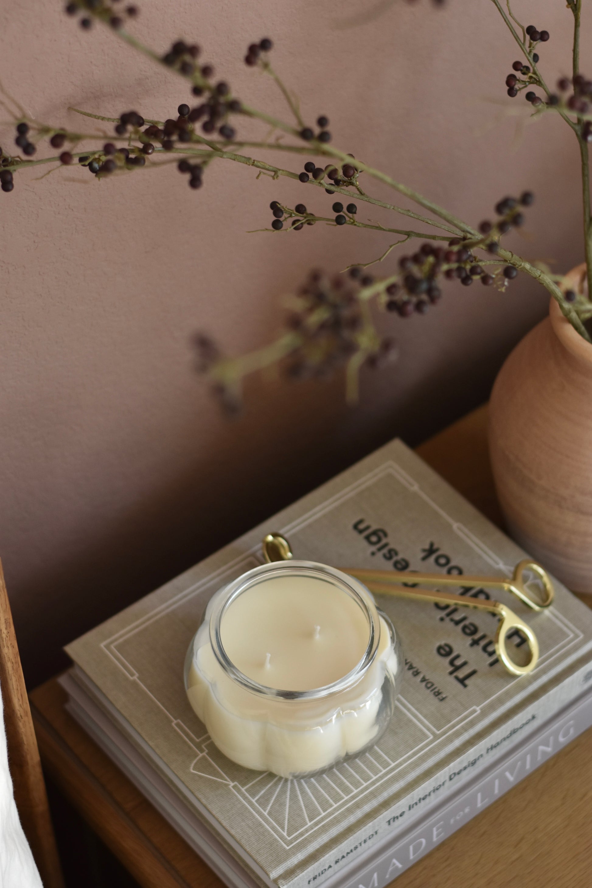 Candle in a decorative container on a book with a pair of wick trimmers, surrounded by dried plants.