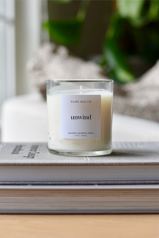Candle labeled 'unwind' by Elsie Mae Co on a stack of books with a blurred background